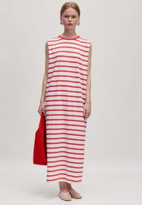 Ärmelloses Maxikleid mit rot-weißem horizontalem Streifenmuster, aus weichem Stoff. Das Model hält eine rote Tasche und trägt transparente Schuhe.