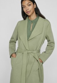 Green wrap coat with a soft texture, oversized collar, long sleeves, and belt detail. Features two front pockets and a relaxed fit.