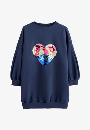 Ragazza giovane in piedi, indossa un abito blu navy oversize con un cuore di paillettes multicolore, leggings blu navy e sneakers alte bianche con fantasie.