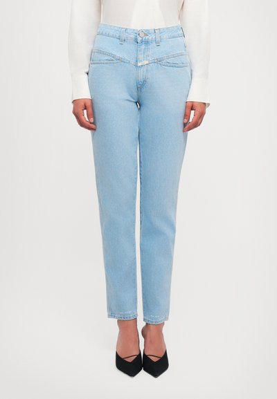 Jean taille haute bleu clair avec une coupe slim, présentant des poches avant et de subtils détails de couture, associée à des chaussures noires à bout pointu.