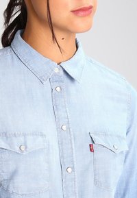 Světle modrá džínová košile s límcem na knoflíky, předními kapsami, stříbrnými knoflíky a malou červenou štítkovou značkou. Měkká textura, ležérní design.