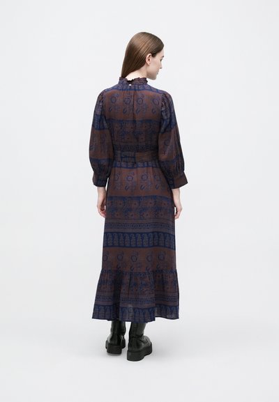 Μάξι φόρεμα σε σκούρο καφέ με μπλε μοτίβα, με φουσκωτά μανίκια, γιακά με βολάν και τελείωμα σε στρώσεις. Φορεμένο με μαύρα μποτάκια στον αστράγαλο.