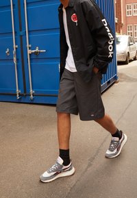 Svart jacka med logo på bröst och ärm, vit t-shirt, grå shorts, grå och vita sneakers med vinröda detaljer, går på asfalt.