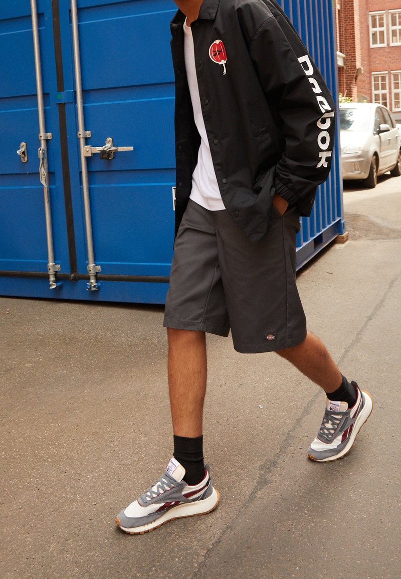 Svart jacka med logo på bröst och ärm, vit t-shirt, grå shorts, grå och vita sneakers med vinröda detaljer, går på asfalt.