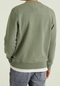 Sweat-shirt vert avec poignets et ourlet côtelés, présentant un panneau arrière texturé et des détails de couture discrets, porté par-dessus une chemise blanche.