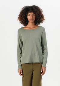 Donna con capelli ricci che indossa una maglietta a maniche lunghe verde salvia e pantaloni verde oliva, in piedi di fronte a uno sfondo bianco uniforme.