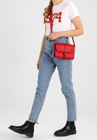 Czerwona torebka na ramię z przednią klapą i metalowym zapięciem, wykonana z gładkiej skóry, noszona z jasno-niebieskimi spodniami z wysokim stanem i czarnymi botkami za kostkę.