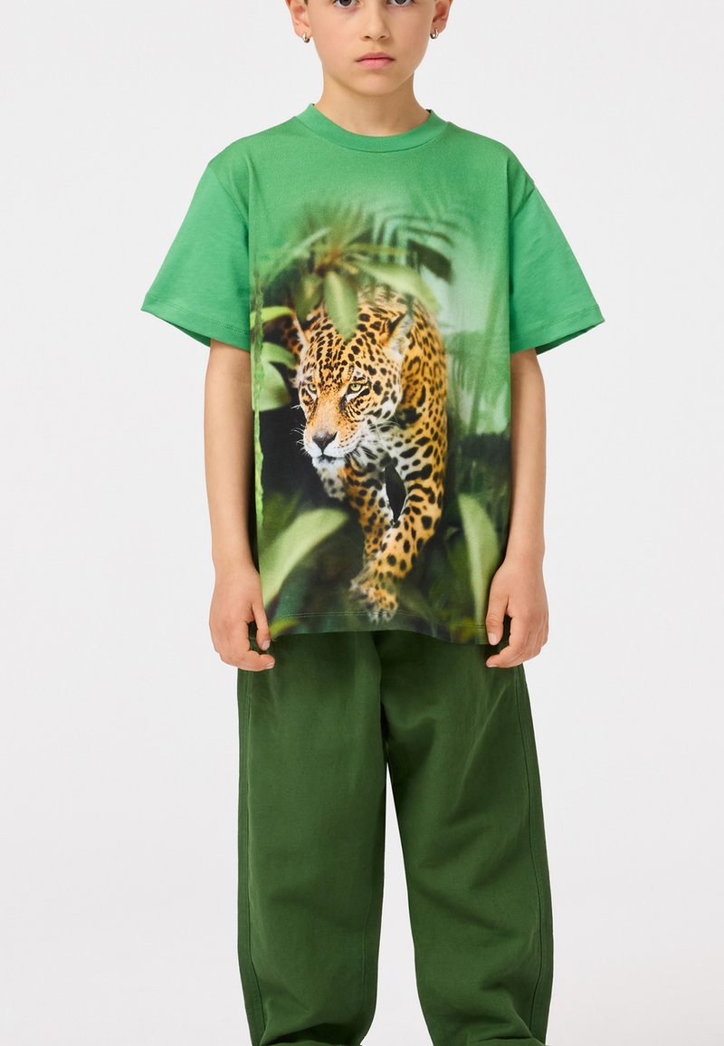 Zelena pamučna majica sa leopard grafikom među lišćem, u kombinaciji s širokim zelenim hlačama, stvara živahnu i opuštenu atmosferu.