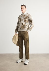 Brown and cream patterned sweater, khaki pants, and light-colored sneakers. Model holds a beige jacket. Neutral background.
