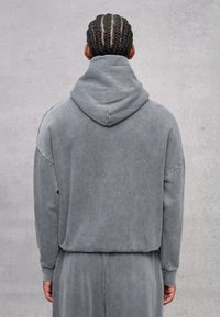 Grau Kapuzenpullover mit weicher Textur, längeren Ärmeln und einem lockeren Schnitt. Sichtbare Nähte entlang der Schultern.
