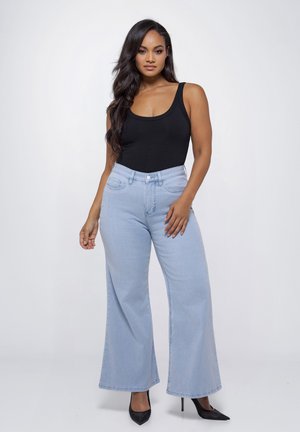 Vrouw met lang donker haar, gekleed in een zwart mouwloos topje, lichtblauwe jeans met wijde pijpen en zwarte hoge hakken, staand tegen een effen achtergrond.