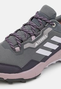 Grauer Sportschuh mit strukturierten Oberflächen, lila Schnürsenkeln, gepolstertem Kragen und weißer Markenkennzeichnung mit drei Streifen an der Seite.