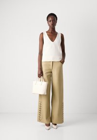Débardeur blanc à col en V sans manches, pantalon beige à jambes larges avec des détails de lacets au bas, tenant un petit sac à main blanc et portant des talons pointus.