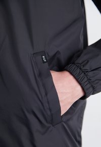 Mustat kevyet takki elastisilla hihansuilla, jossa on sivutasku, jossa on brodeerattu etiketti, jossa lukee "HUF", sileä rakenne ja minimalistinen muotoilu.