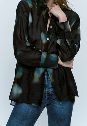 Femme portant une chemise noire ample à boutons avec des taches bleues et beiges, associée à un jean bleu, debout les bras croisés.