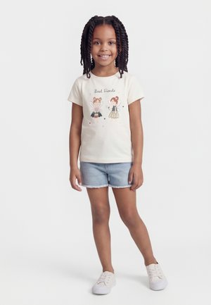 Jeune fille aux cheveux tressés portant un t-shirt blanc "Meilleures amies" et un short en jean, debout devant un fond clair uni, souriante.