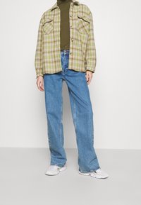 Grünes und beiges karriertes Flanellhemd mit zwei Fronttaschen, kombiniert mit hoch geschnittenen blauen Denim-Jeans und weißen Sneakern.