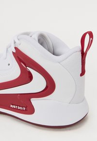 Baskets blanches avec des accents rouges, présentant un design épuré, un matériau texturé et un logo "Just Do It" sur le côté. Forme courbée et languette de traction.