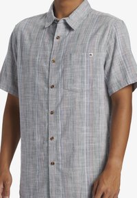 Kurzärmliges Button-Up-Hemd aus leicht grauem und blaugemustertem Stoff. Verfügt über Holzknöpfe und eine Brusttasche mit einem Logopatch.