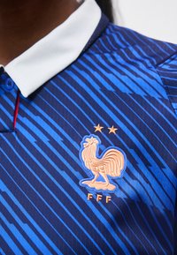 Tricou sportiv cu dungi albastre și albastru închis, cu guler alb, având un emblemă aurie cu cocoș și două stele deasupra literelor „FFF.”