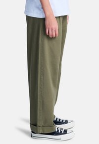 Pantaloni larghi color verde oliva in cotone, con una texture liscia, tasche laterali e abbinati a sneakers in tela bianca e nera.