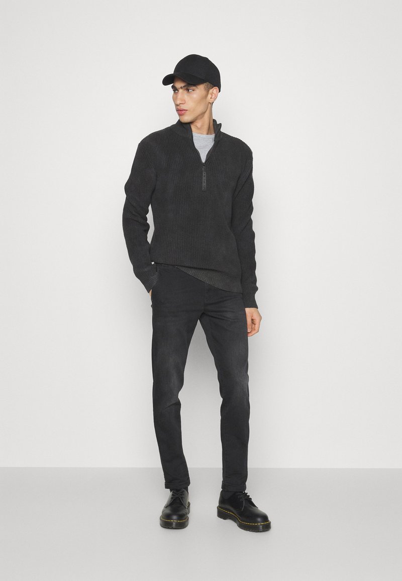 Junger Mann mit schwarzem Baseballcap, dunkelgrauem Zip-up-Pullover, schwarzen Jeans und schwarzen Schuhen, der vor einem schlichten hellen Hintergrund steht.