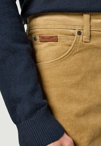 Pantaloni in velluto a coste marrone con una superficie testurizzata, dotati di un'etichetta in pelle e una manica di un maglione blu scuro a maglia parzialmente visibile.