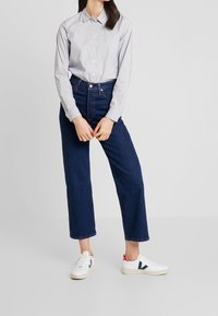 Camicia grigia chiaro con piccoli pois blu, indossata con jeans in denim blu scuro a vita alta e sneaker bianche con accenti neri.