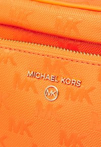 Oranžová textura tašky s pozlaceným zipem, logy Thomas Crown a stříbrným nápisem "MICHAEL KORS". Materiál se zdá být odolný a syntetický.