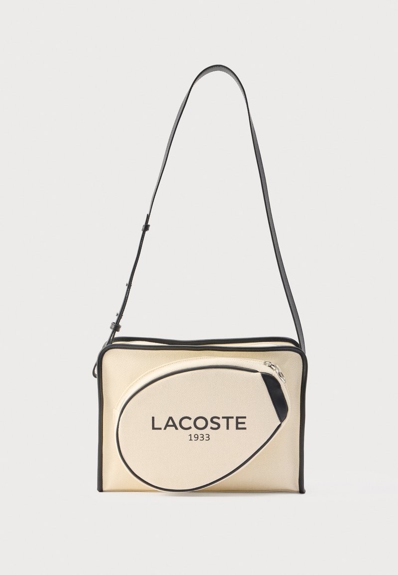 Sac bandoulière en tissu beige texturé avec des accents noirs, doté d'une fermeture éclair et d'une grande poche avant affichant le logo "LACOSTE 1933".