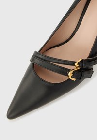 Chaussure à talon haut en cuir noir avec un bout pointu, dotée de deux brides croisées ajustables et de boucles en ton or. Texture lisse et intérieur neutre.