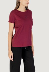 T-shirt di cotone bordeaux con collo a giro, maniche corte e un piccolo dettaglio logo decorativo sul petto. Abbinato a pantaloni neri.