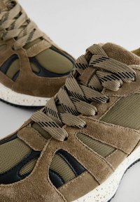 Sneakers mają połączenie oliwkowej siateczki i brązowego zamszu z czarnymi akcentami, teksturowanymi sznurowadłami i nakrapianą białą podeszwą środkową.