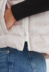 Hellrosa gepolsterte Jacke mit weißem Reißverschluss, gerippten schwarzen Bündchen und einem runden Logo-Patch. Kombiniert mit blauen Denim-Jeans.