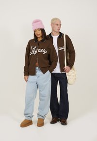 Gilet en tricot marron avec le texte blanc "Legacy", détail de col noir. Jean bleu clair ample, chaussures marron et bonnet en peluche rose.