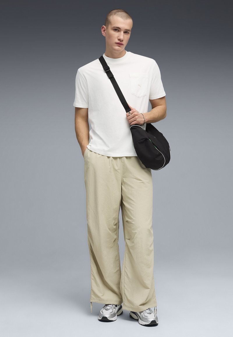 Jeune homme en t-shirt blanc, pantalon large beige, baskets argentées et sac bandoulière noir, posant devant un fond gris dégradé.