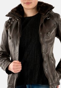 Veste en cuir marron avec un col doublé de fourrure, deux poches poitrine zippées et une coupe ajustée. En dessous, un pull noir côtelé est visible.