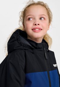 Veste noire et bleue avec une capuche, dotée d'une fermeture éclair et d'un logo sur la poitrine ; texture lisse, design décontracté, sans motifs visibles.