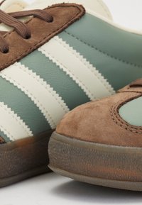 Zapatillas deportivas de cuero verde y crema con detalles en gamuza marrón y suelas texturizadas, que presentan tres rayas blancas y un diseño de cordones.