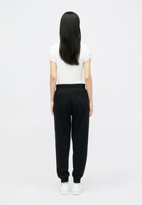 Sieviete stāv, pagriezusies projām, tērpta baltā īsām piedurknēm T-kreklā, melnos trenējošajos biksēs un baltās sporta apavos uz vienkrāsaina baltā fona.