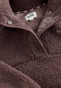 Primo piano di una giacca in pile marrone scuro con texture, bottoni a pressione e un'etichetta bianca con la scritta "self." all'interno del colletto.