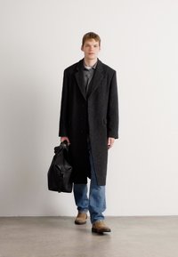 Schwarzer Wollübermantel mit einfacher Knopfreihe, graues Hemd, blaue Jeans und braune Schuhe, hält eine schwarze Lederhandtasche.