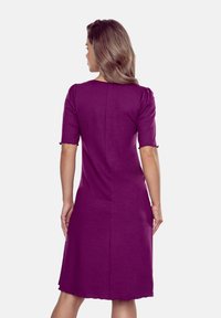 Kobieta widziana jest z tyłu, ubrana w dopasowaną fioletową sukienkę z krótkimi bufkami, z okrągłym dekoltem i długością do kolan.