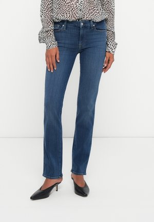 Kobieta w niebieskich prostych dżinsach, czarnych czółenkach na obcasie z ostrym noskiem oraz czarno-białej bluzce w długi rękaw z wzorem.