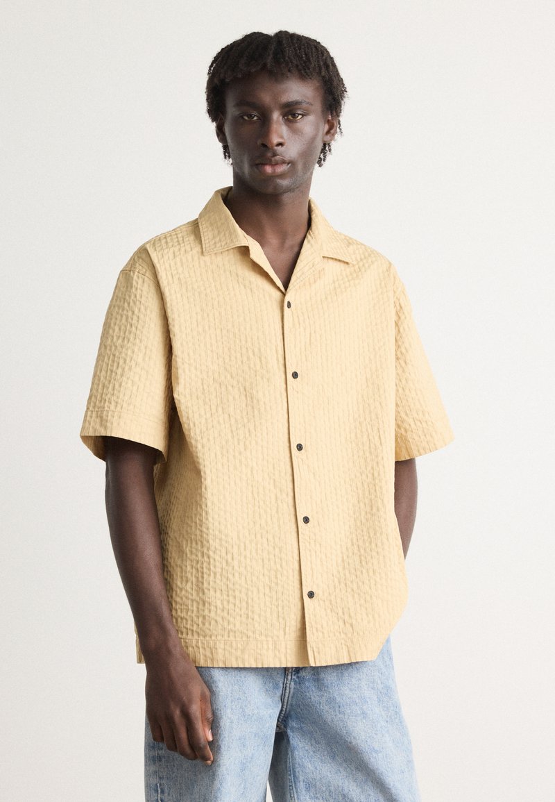 Short-sleeve, textured shirt in pale yellow with a button-down front and black buttons. Paired with light blue denim jeans.