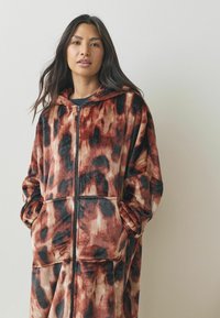 Fleece hooded onesie in warme tinten van bruin en zwart, met een rits en twee voorzakken, gemaakt van een zachte, gestructureerde stof.