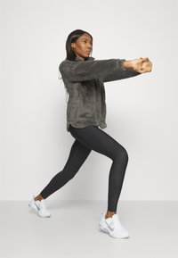 Gray, plush zip-up jacket, black patterned leggings, and white athletic shoes worn by a person in a lunging position against a plain background.