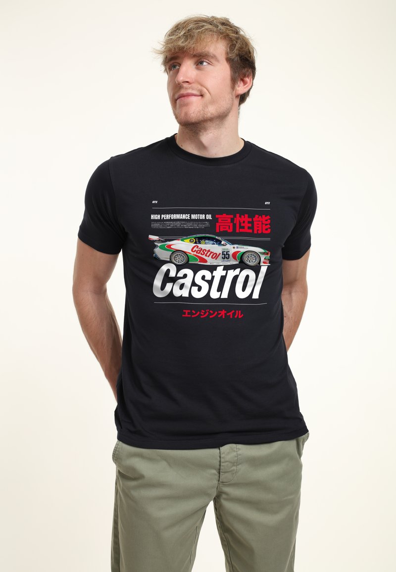 Jeune homme portant un t-shirt noir avec un graphique de voiture de course Castrol, texte rouge et blanc, et un pantalon kaki, debout devant un fond uni.