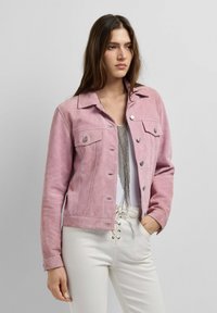 Donna che indossa una giacca di suede rosa con bottoni sopra una maglietta bianca con dettagli in frange metalliche, abbinata a pantaloni bianchi con lacci, in piedi contro uno sfondo neutro.