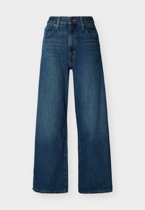 Jean en denim bleu à taille haute et à jambes larges, avec fermeture à boutons et fermeture éclair à l'avant, passants pour ceinture et poches avant sur un fond uni.
